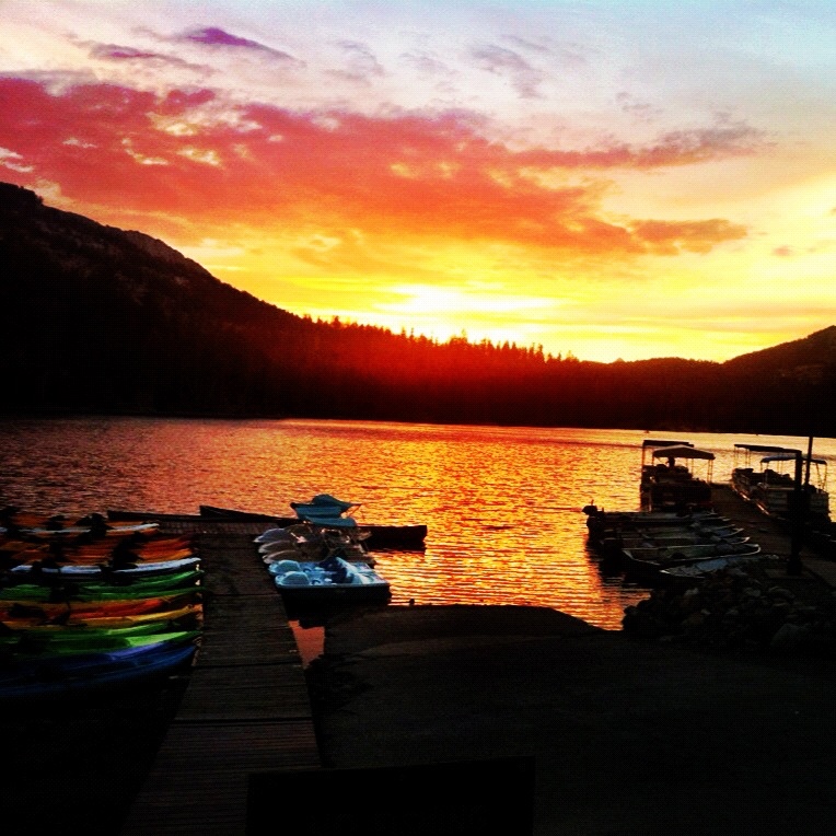 A photo of a brilliant yellow and orange sunset over the lake and mountains with two docks with boats in the foreground