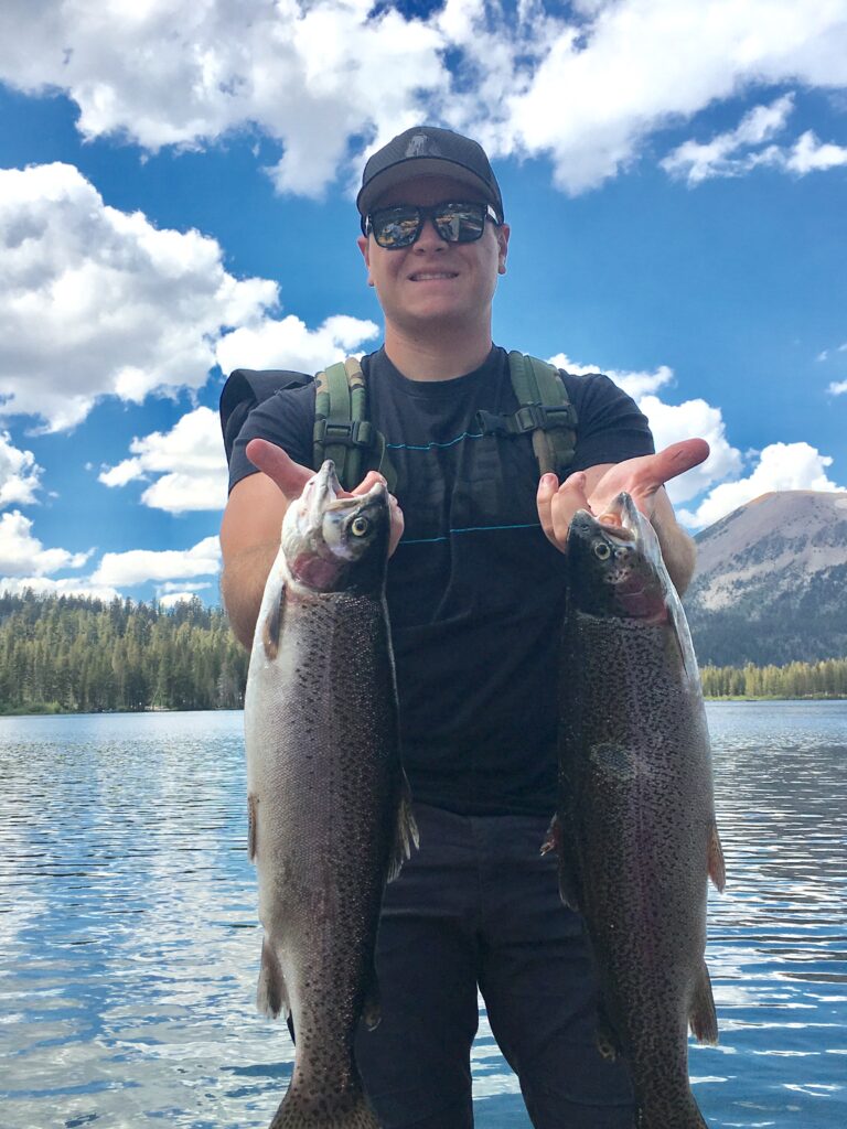 A man holding two rainbow trout with the lake and mountains behind him