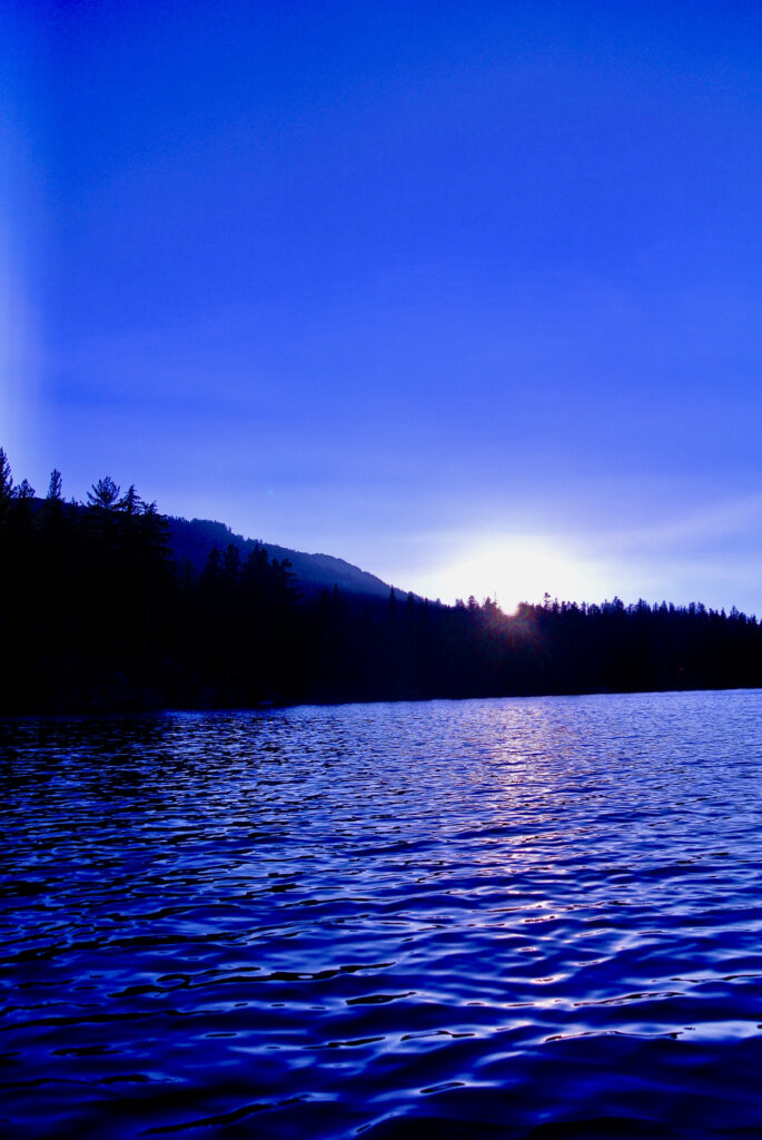 A photo of the sun descending behind the trees and reflecting on the blue lake waters