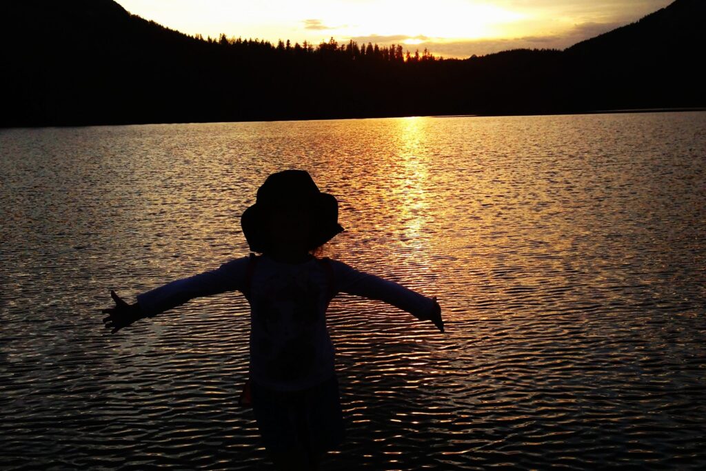 Silhouette of a child standing on the end of the dock with arms outstretched. 