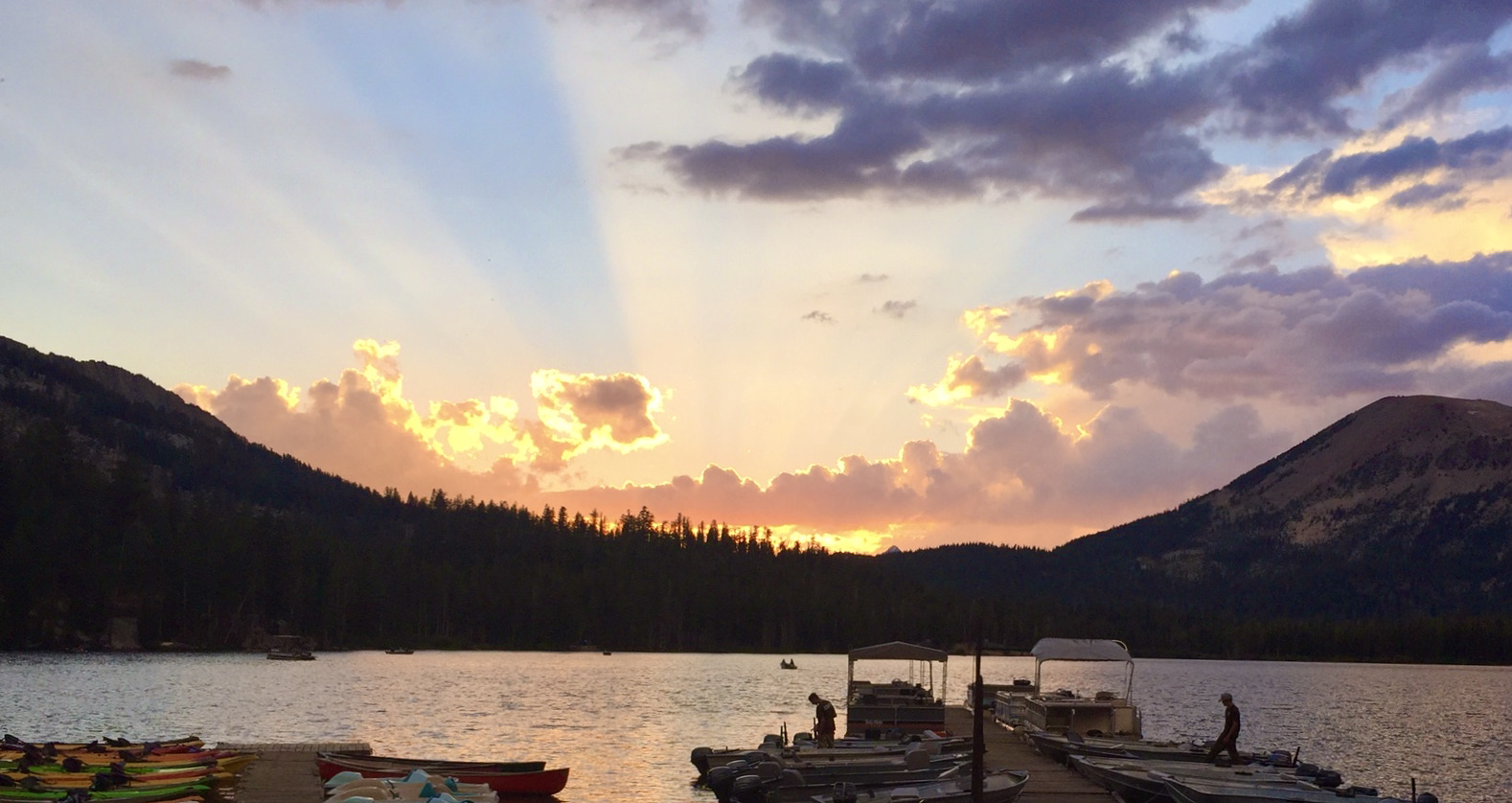 Photo of a sunset with sun rays coming out over the mountains withe boats on the dock in the foreground